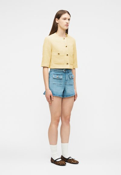 Une femme se tient debout, portant une veste jaune à manches courtes avec des boutons dorés, un short en denim taille haute avec des poches avant, des chaussettes blanches et des ballerines à imprimé léopard.