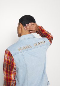 Light blue denim vest with embroidered "KARL KANI" on the back, featuring plaid long sleeves in warm tones of red, orange, and brown.