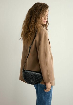 Kobieta o falowanych włosach, ubrana w camelowy płaszcz i niebieskie dżinsy, z czarną, półokrągłą torbą na ramię o fakturze krokodyla przewieszoną przez ramię.