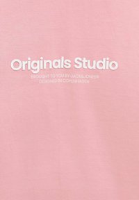 Roza bombažna majica s poudarjenim belim besedilom: "Originals Studio," z manjšim besedilom spodaj, ki označuje blagovno znamko in izvor oblikovanja.