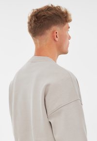 Sudadera de color gris claro con un corte relajado, cuello acanalado, hombros caídos y una textura suave sin logotipos ni patrones visibles.