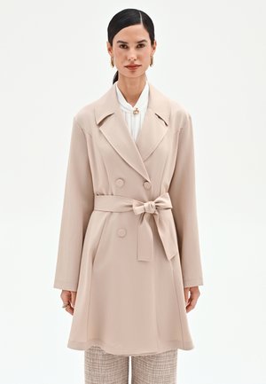 Donna che indossa un trench color beige con cintura e doppi bottoni sopra una camicetta bianca e pantaloni a quadri, con orecchini a cerchio dorati e anelli.