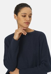 Marineblauer Pullover aus strukturiertem Stoff, mit Rundhalsausschnitt und gerippten Bündchen, getragen von einer Person, die ihre Hand nahe am Kinn hat.