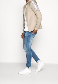 Beige zip-up hoodie med silveraxel detaljer, kombinerat med ljusblå slim-fit jeans och vita sneakers. En enkel, avslappnad outfit.