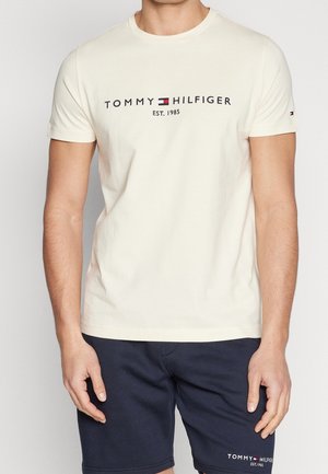 Homme portant un t-shirt crème Tommy Hilfiger avec logo et un short bleu marine, debout devant un fond uni.