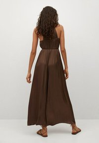 Vestido maxi marrón con plisados y diseño de un solo hombro, textura suave y tela fluida, combinado con sandalias de tiras.