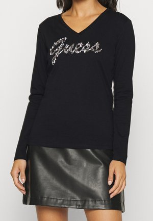 Femme portant un haut noir à manches longues avec un motif à sequins "Grace" et une jupe en cuir noire, debout devant un fond uni.