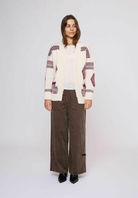 Donna in piedi, che indossa un cardigan a righe bianche e maro, una camicia bianca, pantaloni larghi marroni e scarpe nere a punta, su uno sfondo neutro.