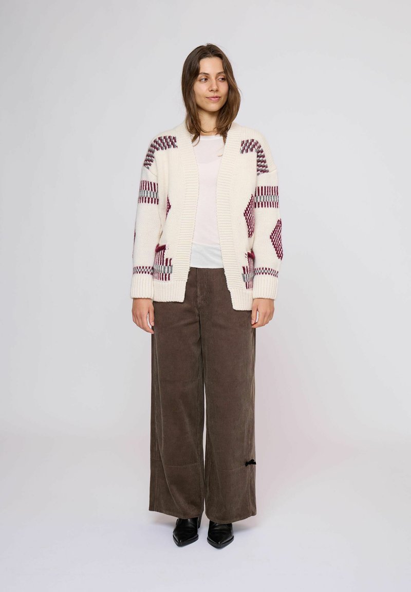 Donna in piedi, che indossa un cardigan a righe bianche e maro, una camicia bianca, pantaloni larghi marroni e scarpe nere a punta, su uno sfondo neutro.
