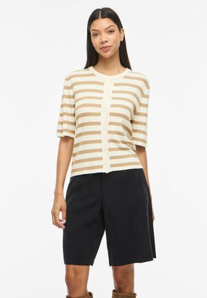 Kvinne med langt mørkt hår som har på seg en beige og kremfarget stripete kortermet knappet genser og svarte shorts til knærne.