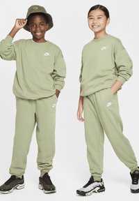 Zöld Nike pulóverek fehér logóval, passzoló zöld melegítőnadrágokkal. Az egyik gyerek zöld vödörkalapot visel; mindketten különböző fekete sportcipőt hordanak.