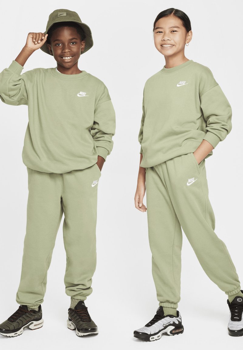 Zöld Nike pulóverek fehér logóval, passzoló zöld melegítőnadrágokkal. Az egyik gyerek zöld vödörkalapot visel; mindketten különböző fekete sportcipőt hordanak.