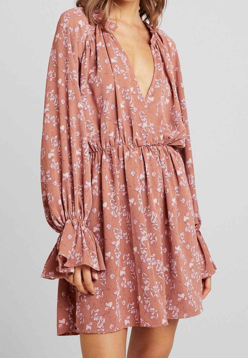 Femme portant une robe fleurie ample, à manches longues, rose poudré, avec un profond décolleté en V et une taille élastique froncée.