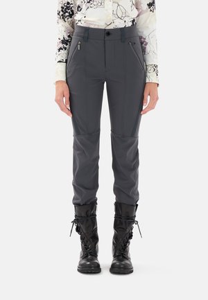 Donna che indossa pantaloni aderenti grigio scuro con tasche frontali con zip, una camicia a fiori con bottoni e stivali da combattimento neri con lacci.