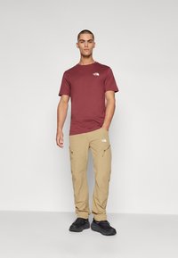Burgunder T-skjorte og beige cargo-bukser, begge laget av stoff, med logo-detaljer. Bukser har lommer og en avslappet passform. Svarte sneakers fullfører antrekket.