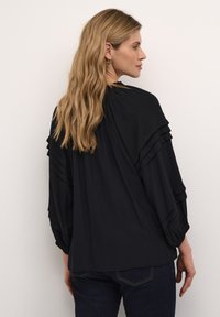Blusa nera con dettagli arricciati lungo le spalle e le maniche. Tessuto morbido con una vestibilità ampia, abbinata a jeans scuri.