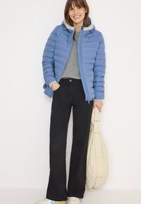 Veste matelassée bleu clair avec un sweat à capuche gris en dessous, pull gris, pantalon noir et baskets blanches. Porte un sac à bandoulière crème.