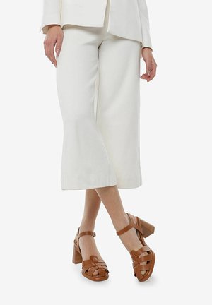 Sandalias de tacón cuadrado de cuero marrón con un diseño de tiras cruzadas. Cuentan con un tacón resistente y una correa para el tobillo, combinadas con unos pantalones de pierna ancha color crema.