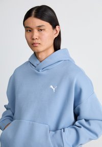 Lysblå hættetrøje lavet af blødt stof, med en frontlomme og et hvidt Puma-logo på brystet. Glat tekstur, afslappet pasform.