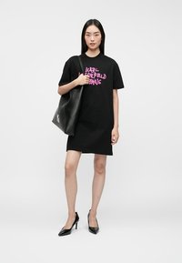 Czarna sukienka t-shirt z różowym graficznym logo, w połączeniu z dużą czarną torbą na ramię. Modelka nosi czarne szpilki. Gładka tekstura materiału.