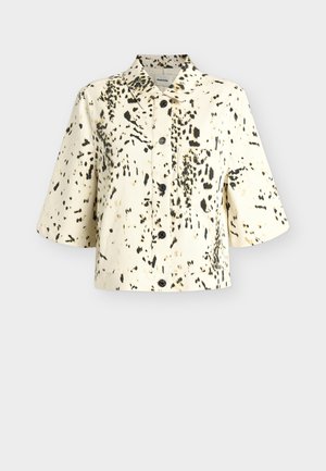 Kurzärmelige cremefarbene Jacke mit schwarzen unregelmäßigen Flecken, durchgeknöpfter Vorderseite und spitzem Kragen auf schlichtem hellem Hintergrund.