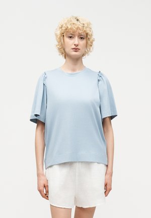 Osoba o krótkich kręconych blond włosach nosi jasnoniebieską bluzkę z krótkim rękawem z marszczonymi ramionami i białe szorty na jednolitym tle.