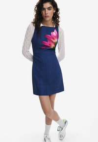 Rochie albastră din denim, cu o formă ajustată, având un imprimeu mare cu flori roz pe față. Purtață peste un top alb transparent, cu textură, cu mâneci lungi.