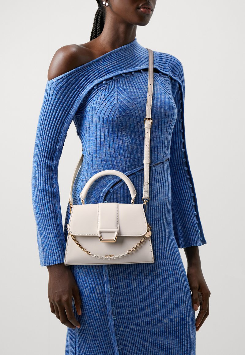 Petit sac à main beige avec un design structuré, doté d'un accent chaîne et d'une anse supérieure. Porté en bandoulière avec une robe bleue côtelée texturée.