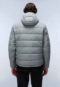 Mand med krøllet hår iført en lys grå quiltet pufferjakke med hætte, der står med ryggen til mod en simpel lys baggrund.