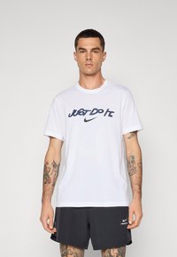 White cotton t-shirt featuring a navy "JUST DO IT." logo and Nike swoosh. The model wears black shorts, showcasing arm tattoos.