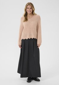 Maglione a maglia rosa chiaro con scollo a V e orlo scallop, abbinato a una lunga gonna nera plissettata e scarpe nere eleganti.