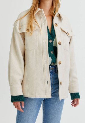 Femme portant une veste en laine crème boutonnée sur un cardigan vert à motifs et un jean bleu, debout, les bras détendus le long du corps.