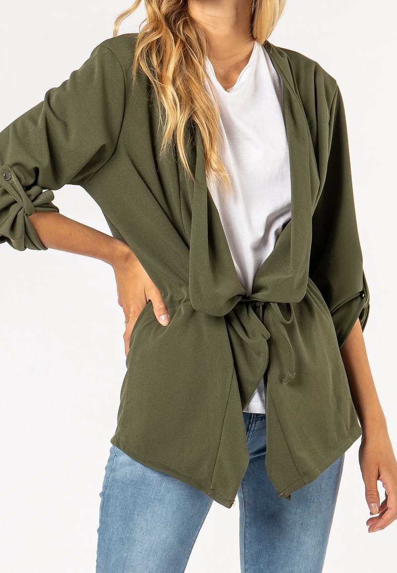 Femme portant une veste ouverte couleur olive avec des manches retroussées nouées à la taille, chemise blanche et jean bleu.