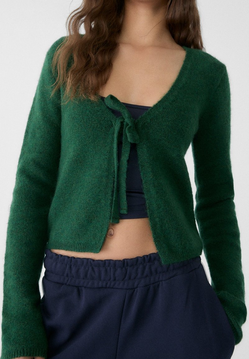 Žena v tmavě zeleném pleteném kardiganu uvázaném přes černý crop top a námořnicky modrých elastických kalhotách.