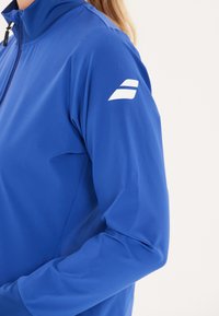 Blå atletisk jakke med høj krave, lynlåsgylpter foran og glat tekstur. Har et hvidt logo på venstre ærme.