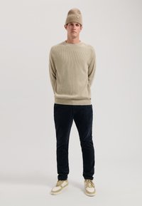Beiger Rippstrickpullover, marineblaue Cordhose, cremefarbene und beige Sneakers; Model trägt eine passende Mütze mit Logodetail.