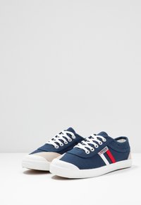 Marineblå lærreds sneakers med hvide snørebånd og gummisåler. Har røde og hvide striber på siden og lysegrå ruskindsdetaljer.