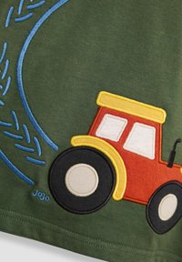 Zöld pamut szövet, amelyen egy hímzett piros traktor látható sárga tetővel és fekete kerekekkel, kék öltésekkel kirakott gumiabroncs nyomokkal kísérve.