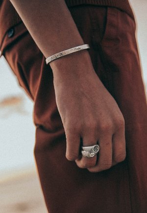 Silver bangle with textured surface and engraved symbols on a wrist, paired with a chunky ring featuring a peace sign design on a hand.