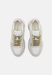 Baskets blanches et beige avec des lacets colorés bleus et jaunes, des accents roses au niveau du talon et une semelle texturée. Logo "GUESS" sur la languette.