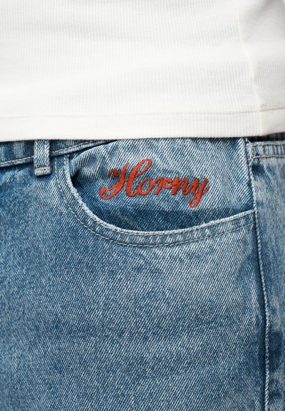 Κοντινό πλάνο τσέπης από μπλε τζιν με κόκκινη κεντημένη λέξη σε καλλιγραφικό στυλ που γράφει «Horny» κάτω από το λευκό υφασμάτινο τελείωμα πουκάμισου.
