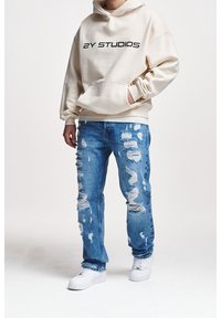 Cremefarbener Hoodie mit schwarzem "2Y STUDIOS"-Schriftzug, kombiniert mit hellblauen, stark zerrissenen Jeans und weißen Turnschuhen.
