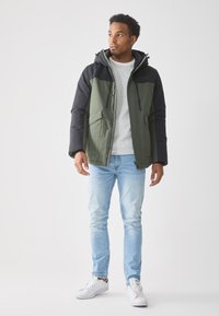 Polstret jakke i olivengrøn og sort, med hætte, to frontlommer og lynlåslommer på brystet. Stylest med lyseblå jeans og hvide sneakers.