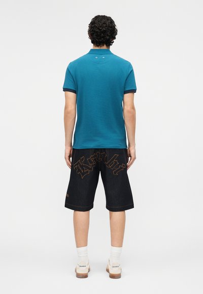 Άνδρας με σγουρά μαλλιά που φοράει τυρκουάζ μπλουζάκι πόλο με κοντά μανίκια, μαύρες βερμούδες μήκους μέχρι το γόνατο με πορτοκαλί γραφικά κείμενα, λευκές κάλτσες και λευκά αθλητικά παπούτσια.