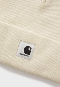 Căciulă tricotată de culoare crem, cu margine pliată, având un eticheta rectangulară albă cu logo-ul Carhartt în negru.