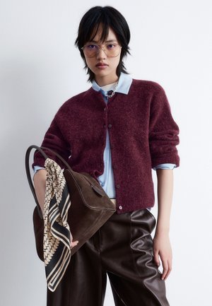 Burgundy knitted cardigan with round buttons over a light blue shirt. Model wears oversized brown leather pants, holding a brown bag with a patterned scarf.