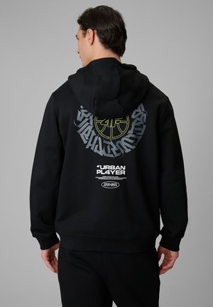 Mężczyzna w czarnej bluzie z kapturem z okrągłym logo sportowym i napisem "URBAN PLAYER" na plecach, stojący na jednolitym szarym tle.