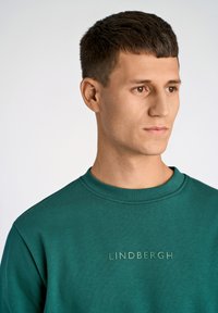 Grön sweatshirt med rund halsringning och texturerad "LINDBERGH"-logotyp i matchande färg. Tillverkad av ett mjukt material och har långa ärmar.