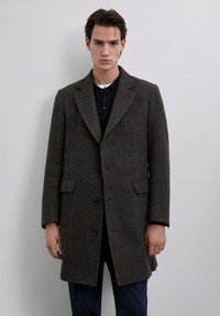 Abrigo de lana marrón oscuro con cuello en pico, dos bolsillos frontales y tres botones negros. Llevado sobre una camiseta polo negra.