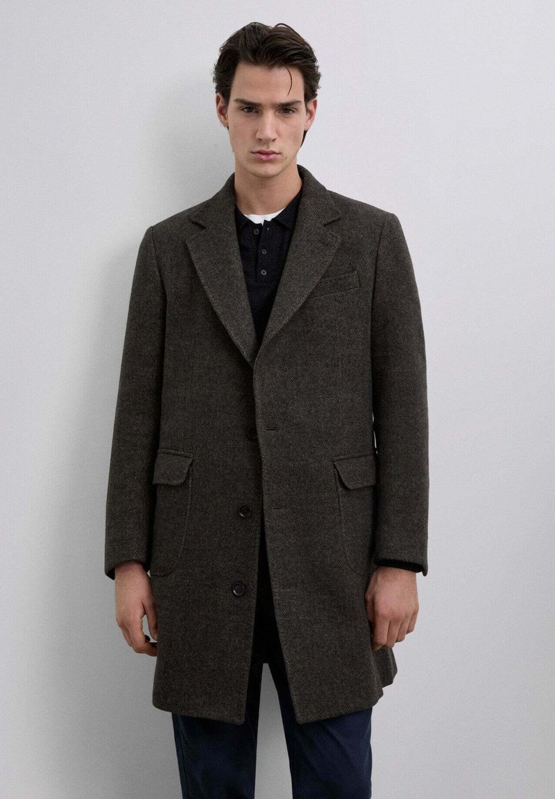 Abrigo de lana marrón oscuro con cuello en pico, dos bolsillos frontales y tres botones negros. Llevado sobre una camiseta polo negra.